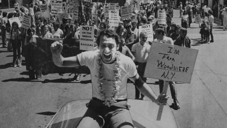 Harvey Milk rides a car at the Gay Pride Parade, leading hundreds of people. He smiles, raises a fist, and holds a sign reading "I'm from Woodmyre NY." Gay Liberation, baby!